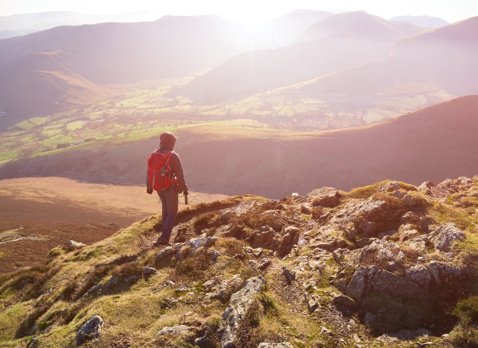 Person with a backpack standing on a rocky mountain ridge overlooking a sunlit valley and distant hills.