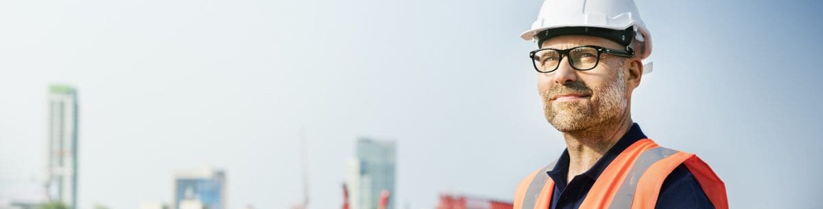 a man wearing glasses, stood wearing a hardhat and an orange PPE vest.