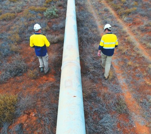Two workers in high‑visibility clothing and hard hats walking alongside an above‑ground pipeline.