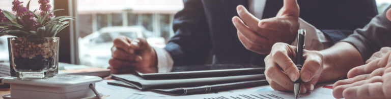 Close‑up of hands holding a pen and discussing printed documents across a desk during a meeting.