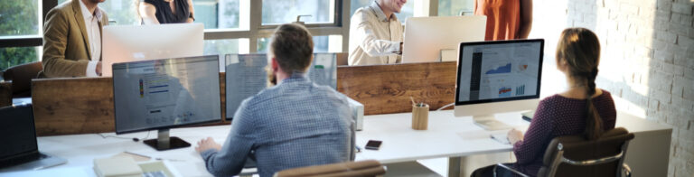 People seated at shared desks using desktop computers and laptops in a bright open‑plan office.