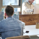 People seated at shared desks using desktop computers and laptops in a bright open‑plan office.