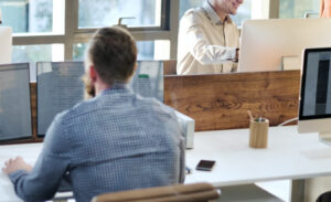 People seated at shared desks using desktop computers and laptops in a bright open‑plan office.