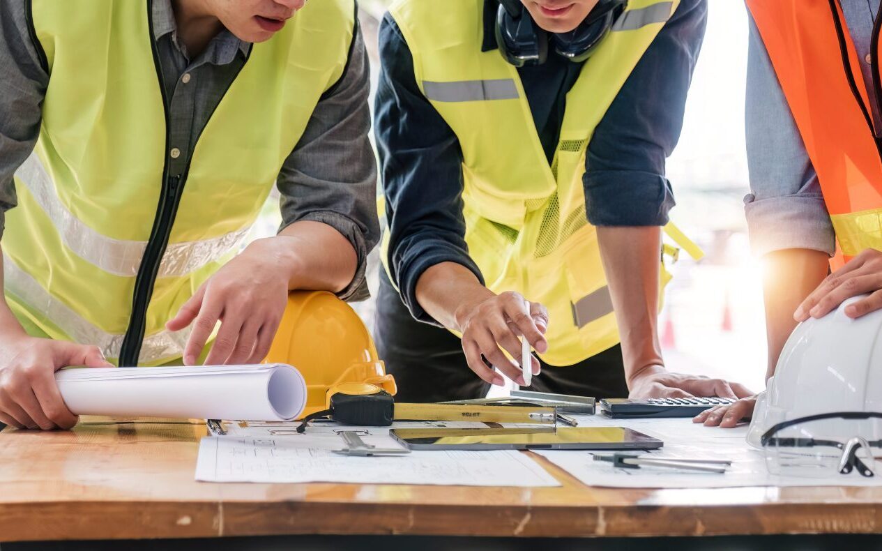Three people in high‑visibility vests reviewing plans, tools and documents on a table indoors.