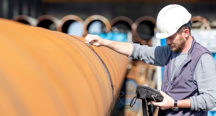 Worker wearing a hard hat inspecting the surface of a large metal pipeline using handheld equipment.