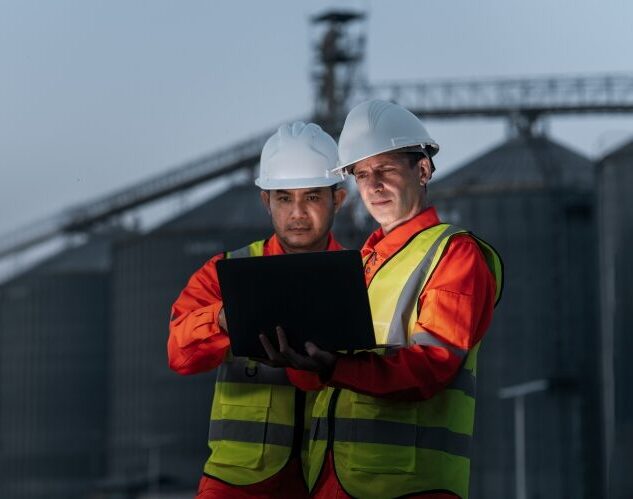 Two workers in high‑visibility clothing and helmets reviewing a tablet at an industrial facility.