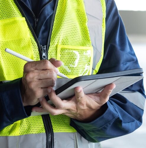Close‑up of a worker in a high‑visibility vest using a stylus on a tablet device indoors.