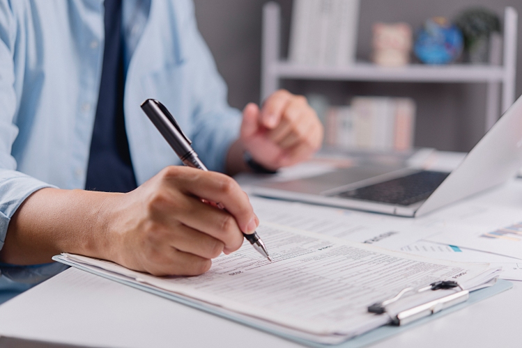 Close‑up of hands using a pen to review and complete paperwork at a desk with a laptop nearby.