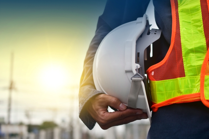 Worker holding a white safety helmet while wearing a high‑visibility vest at an outdoor site.