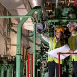 Two workers in high‑visibility vests and hard hats reviewing plans and inspecting green pipework indoors.