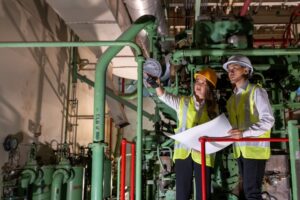 Two workers in high‑visibility vests and hard hats reviewing plans and inspecting green pipework indoors.