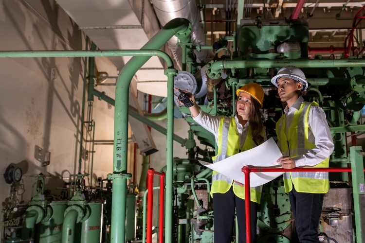 Two workers in high‑visibility vests and hard hats reviewing plans and inspecting green pipework indoors.