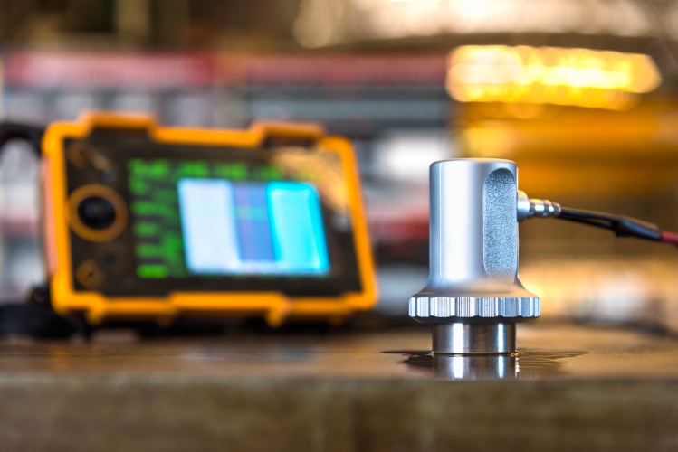 Close‑up of an ultrasonic testing probe on a metal surface with a digital inspection device blurred behind.