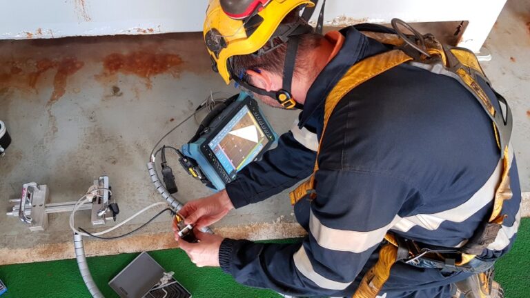 Worker wearing safety gear using handheld testing equipment connected to a digital inspection device indoors.