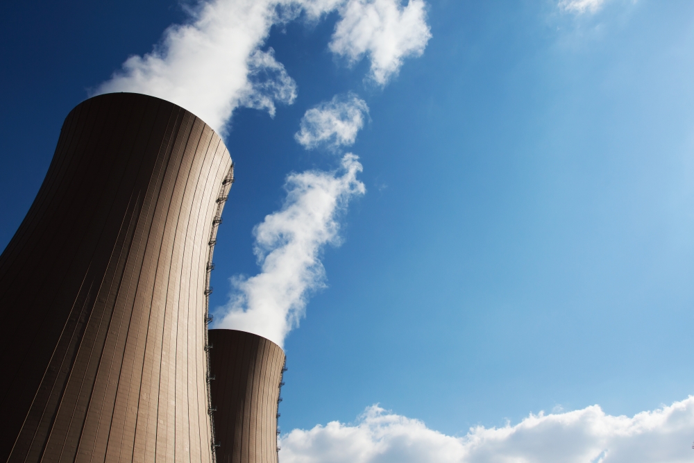 Two large cooling towers releasing white steam into a clear blue sky above an industrial site.