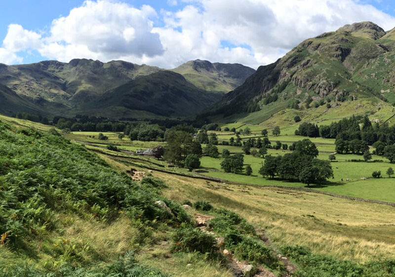Green valley with trees and fields between hills and mountains under a partly cloudy sky.