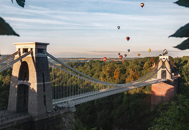 Large suspension bridge over a gorge with hot air balloons floating in the sky above.