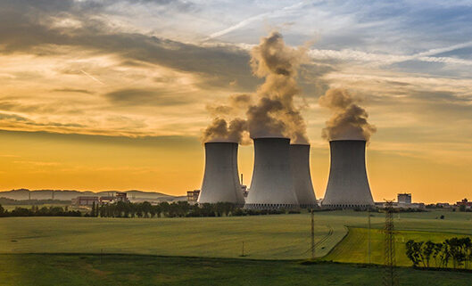 Three large cooling towers release steam above a green field under a cloudy sky.